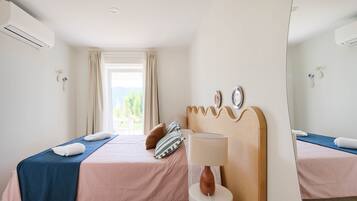 A hotel room with two beds, a window with curtains, a bedside table with a lamp, and a wall-mounted air conditioner.