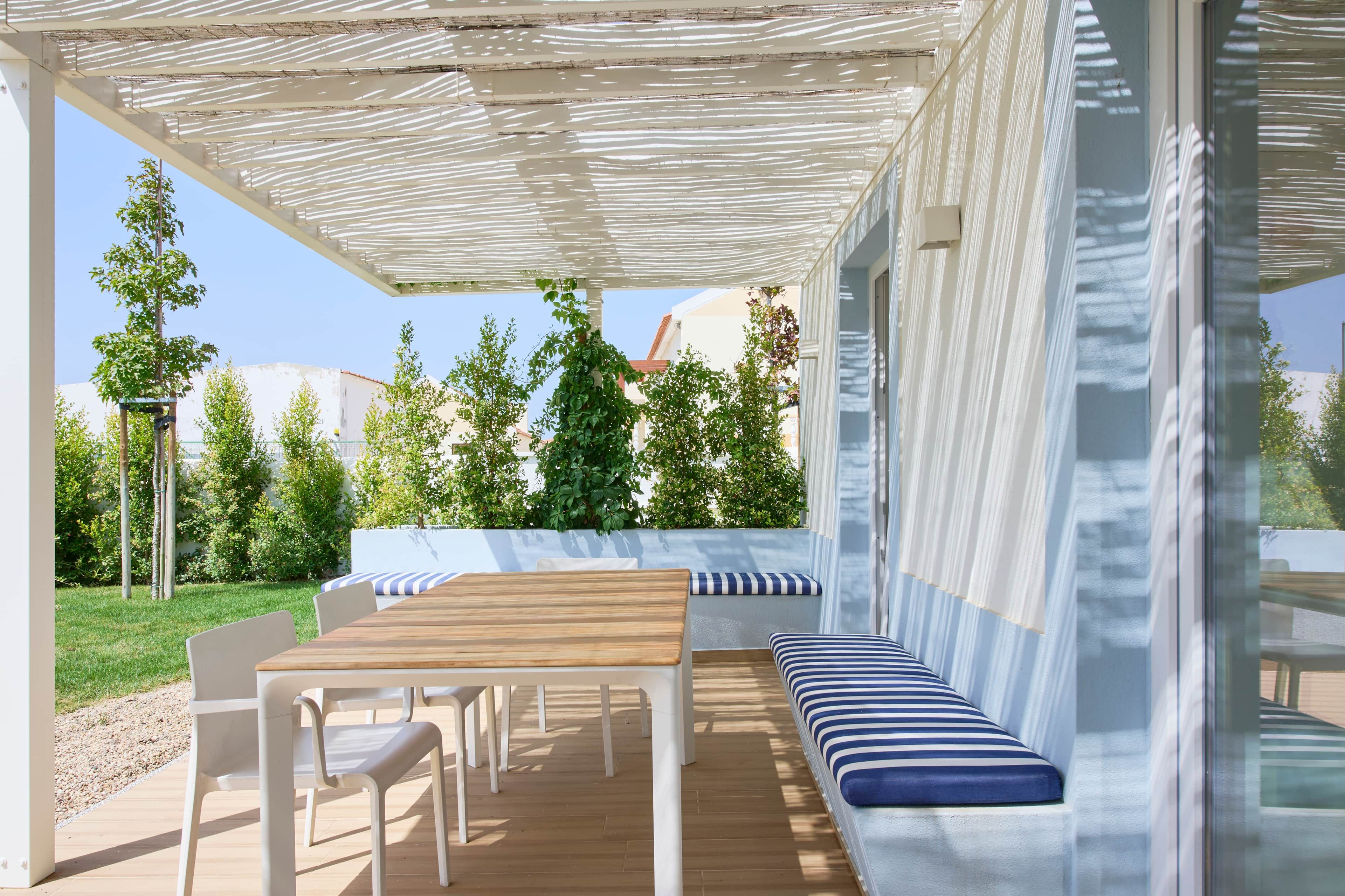 An outdoor patio with a wooden table and chairs, a bench with striped cushions, and a pergola providing shade.