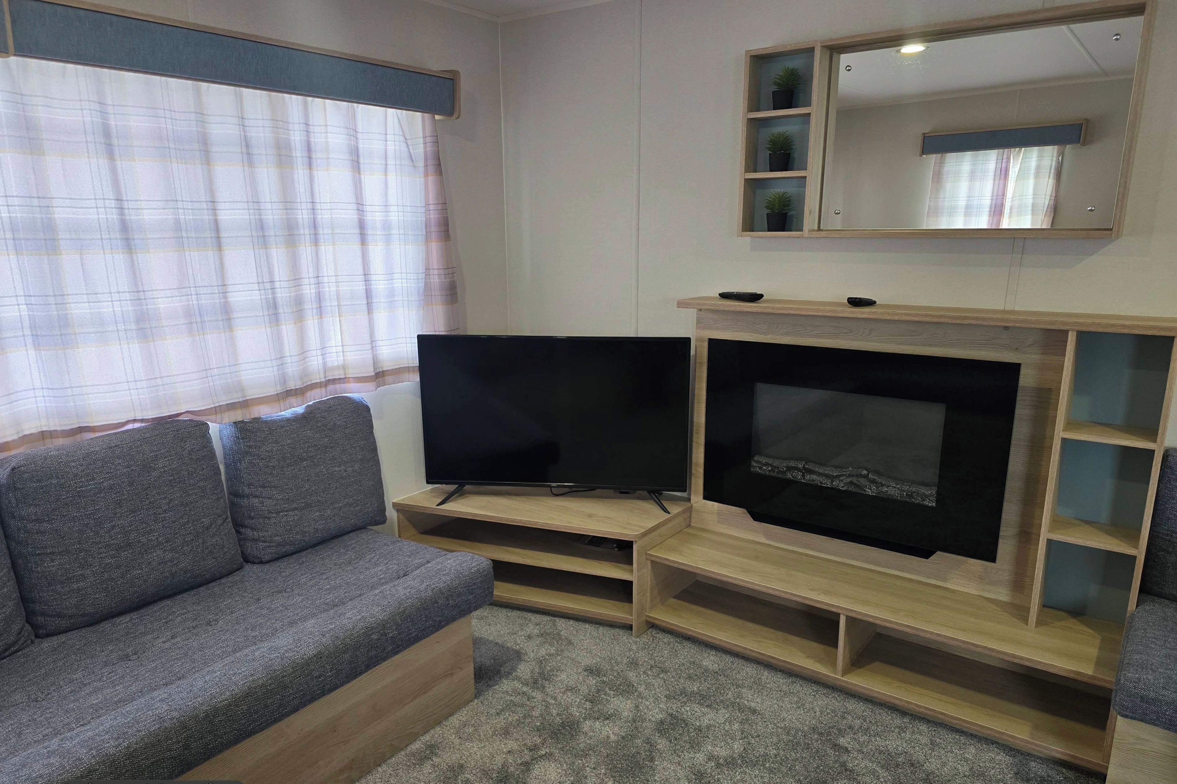 A living room with a grey sofa, a flat-screen TV on a wooden stand, a fireplace, and a window with curtains.