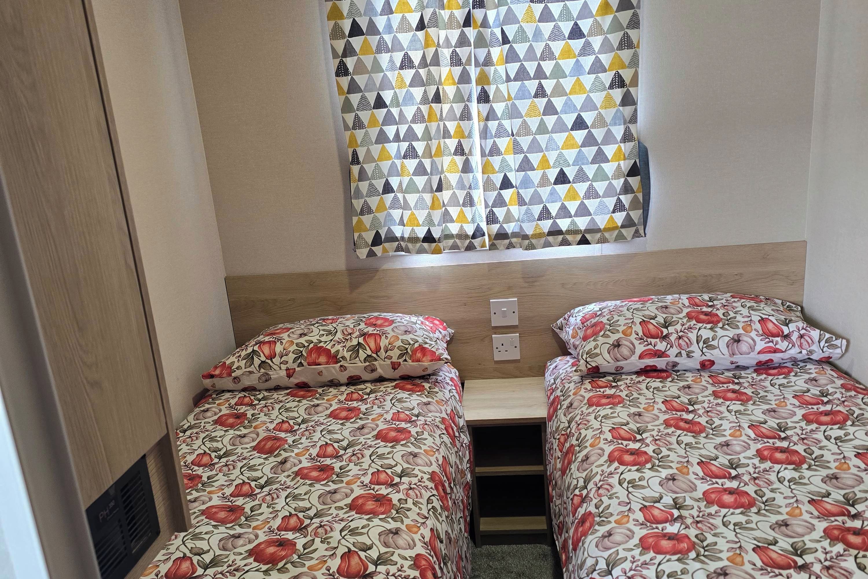 A small, compact room with two beds, a window with patterned curtains, and a wooden headboard.