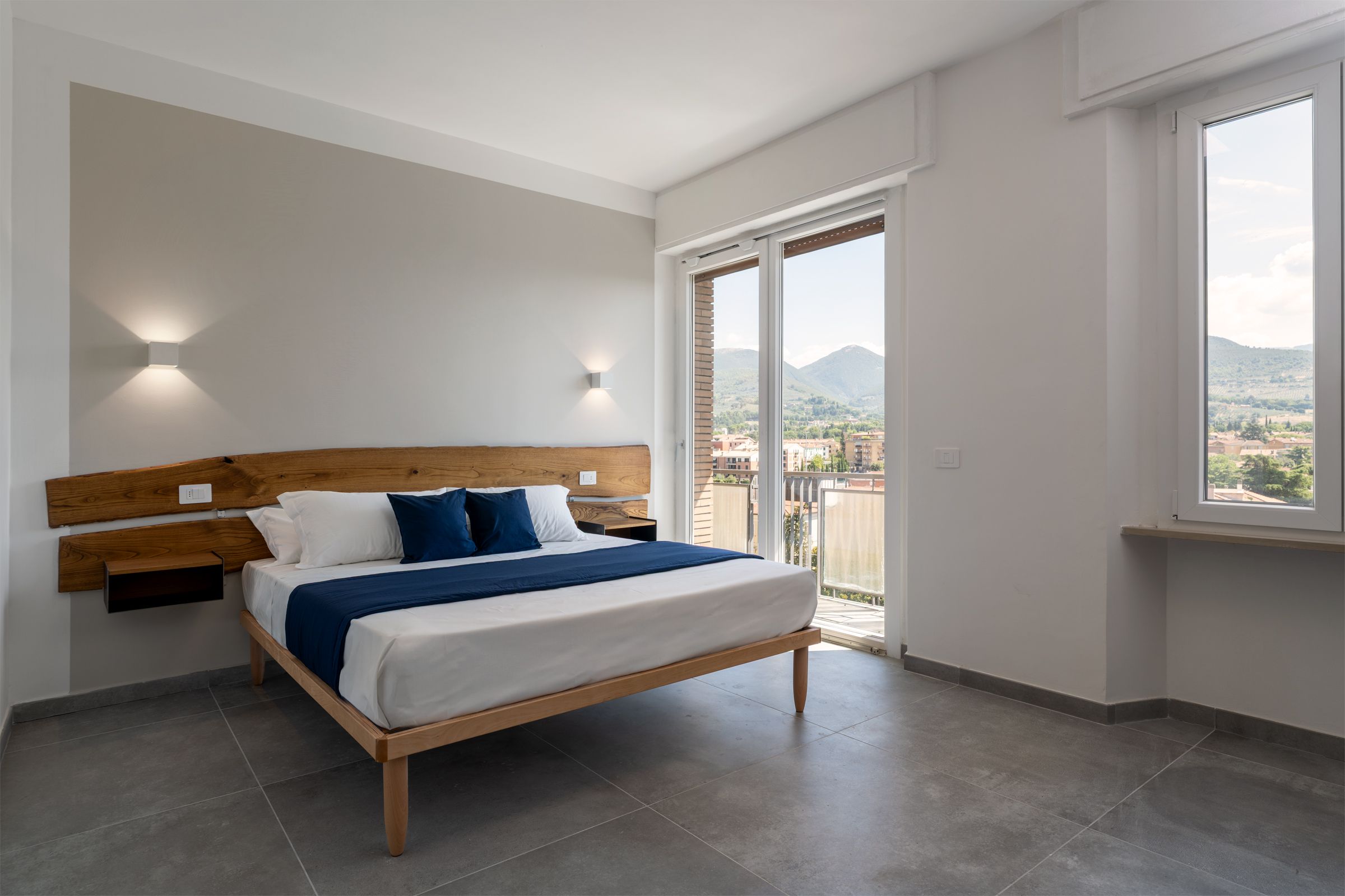Ein Schlafzimmer mit einem Bett, einem Holzkopfende, einem Nachttisch, einer Wandleuchte und einem großen Fenster mit Blick auf Gebäude und Hügel.