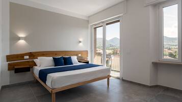Ein Schlafzimmer mit einem Bett, einem Holzkopfende, einem Nachttisch, einer Wandleuchte und einem großen Fenster mit Blick auf Gebäude und Hügel.