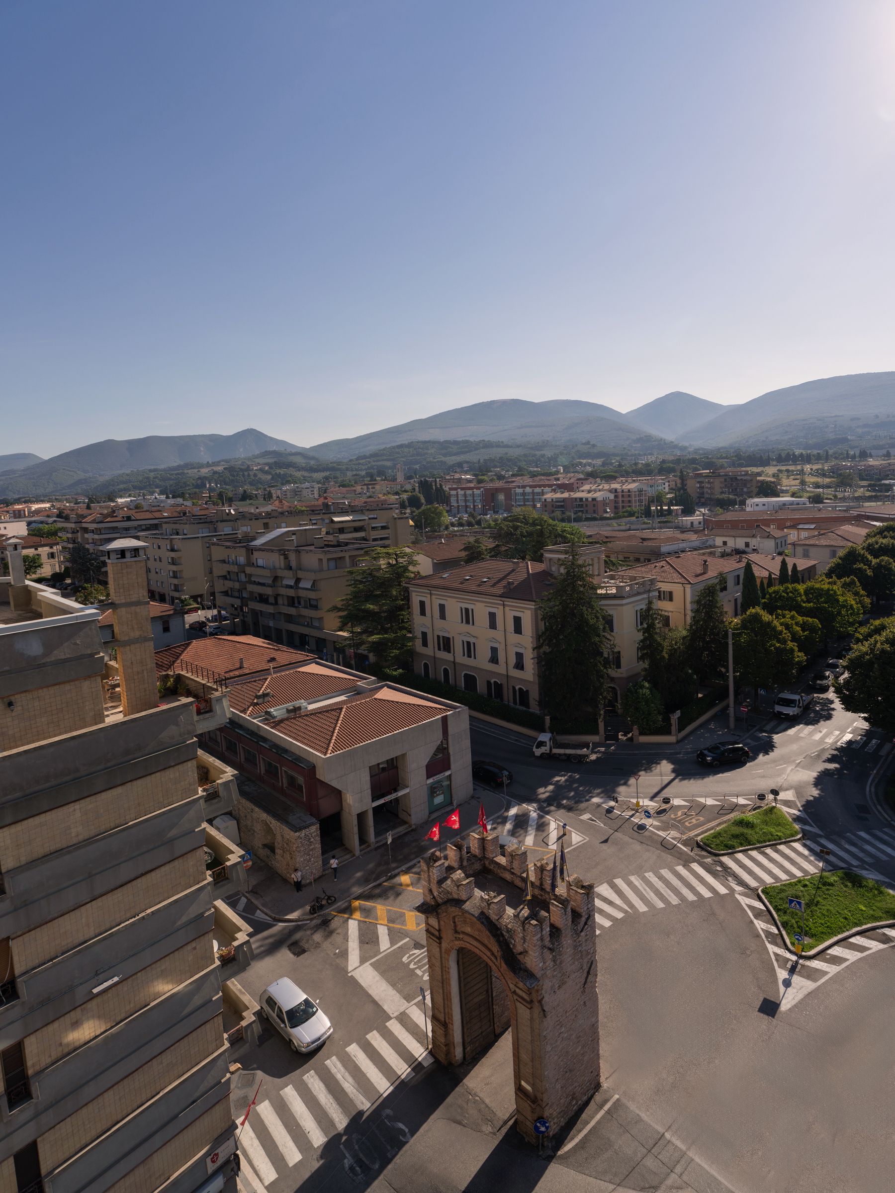 Panoramica urbana con un edificio che spicca per un'ampia arcata, un attraversamento pedonale e una vista chiara della zona circostante e delle montagne in lontananza.