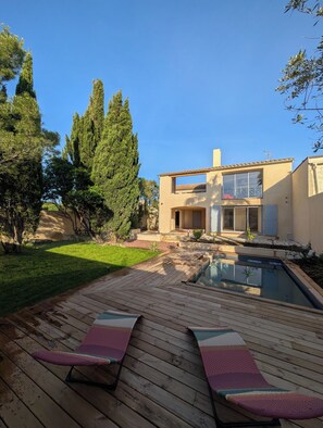 Ein modernes Haus mit Swimmingpool, einer Holzterrasse und Liegestühlen sowie einem gepflegten Rasen.