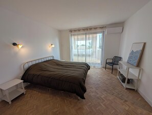Ein Schlafzimmer mit einem Bett, einem Sessel, einem Nachttisch, einem Fenster mit Vorhängen und einem Wandbild.