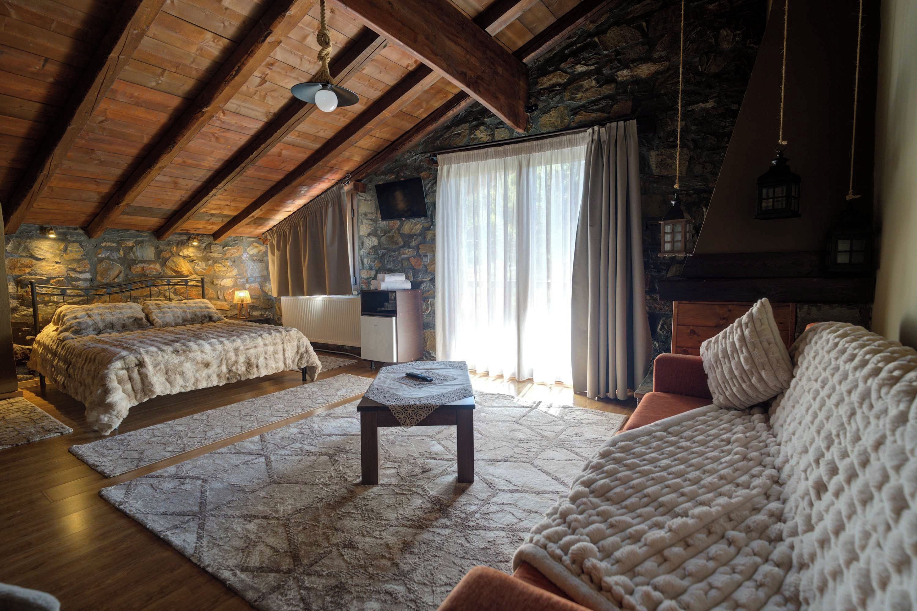 A cozy loft bedroom with a stone wall, a wooden ceiling, and a large window with curtains.