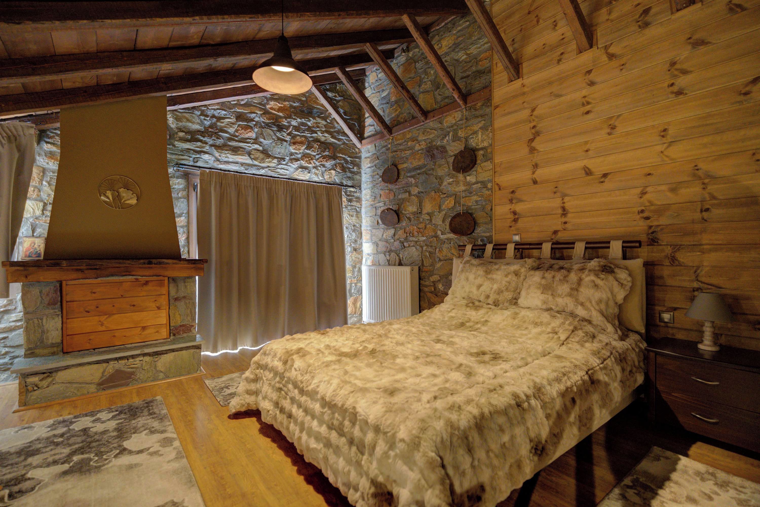 Ein rustikales Schlafzimmer mit Steinwand, Holzbalken, einem Bett mit flauschiger Decke, einer Holzkommode und einer hängenden Leuchte.