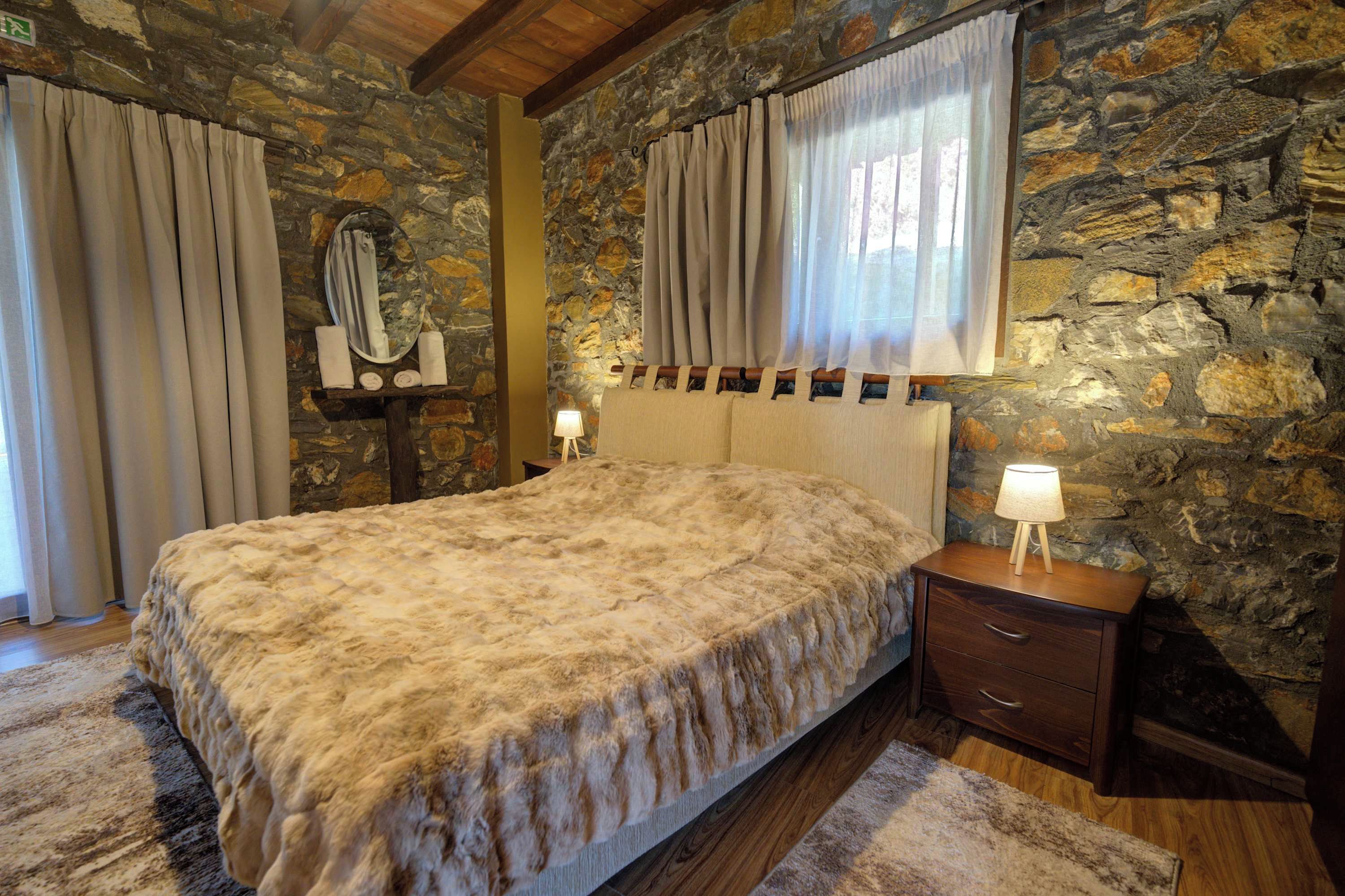 A bedroom with a stone wall, a bed with a fluffy blanket, a nightstand with a lamp, and a window with curtains.