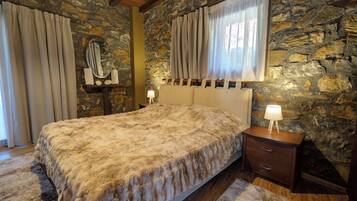 A bedroom with a stone wall, a bed with a fluffy blanket, a nightstand with a lamp, and a window with curtains.