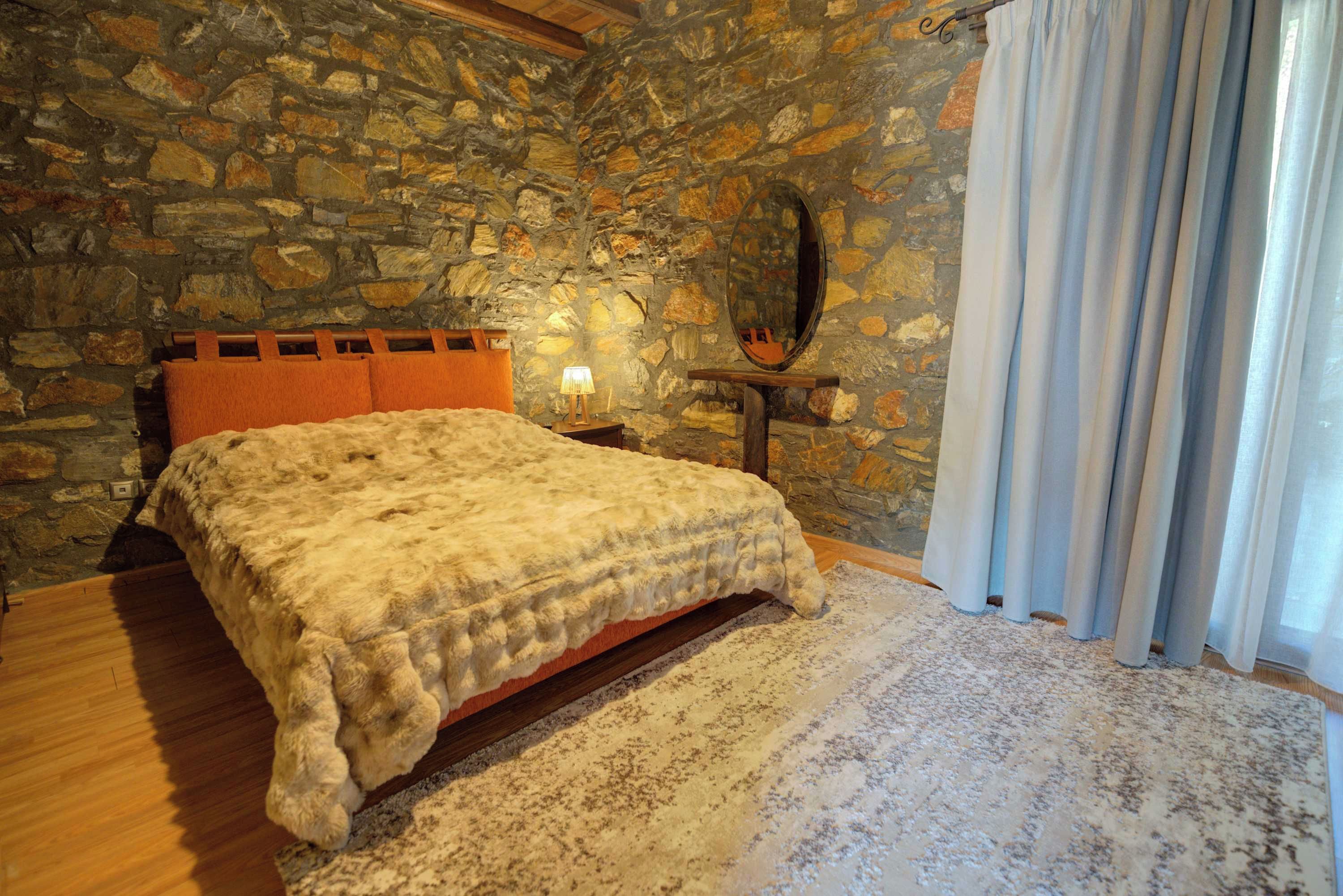 Un dormitorio con pared de piedra, una cama de madera, una mesita de noche con lámpara y una ventana con cortinas.