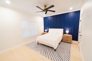 Une chambre à coucher comprenant un lit, deux tables de chevet, un ventilateur de plafond, une fenêtre avec des stores et un tapis à motifs.