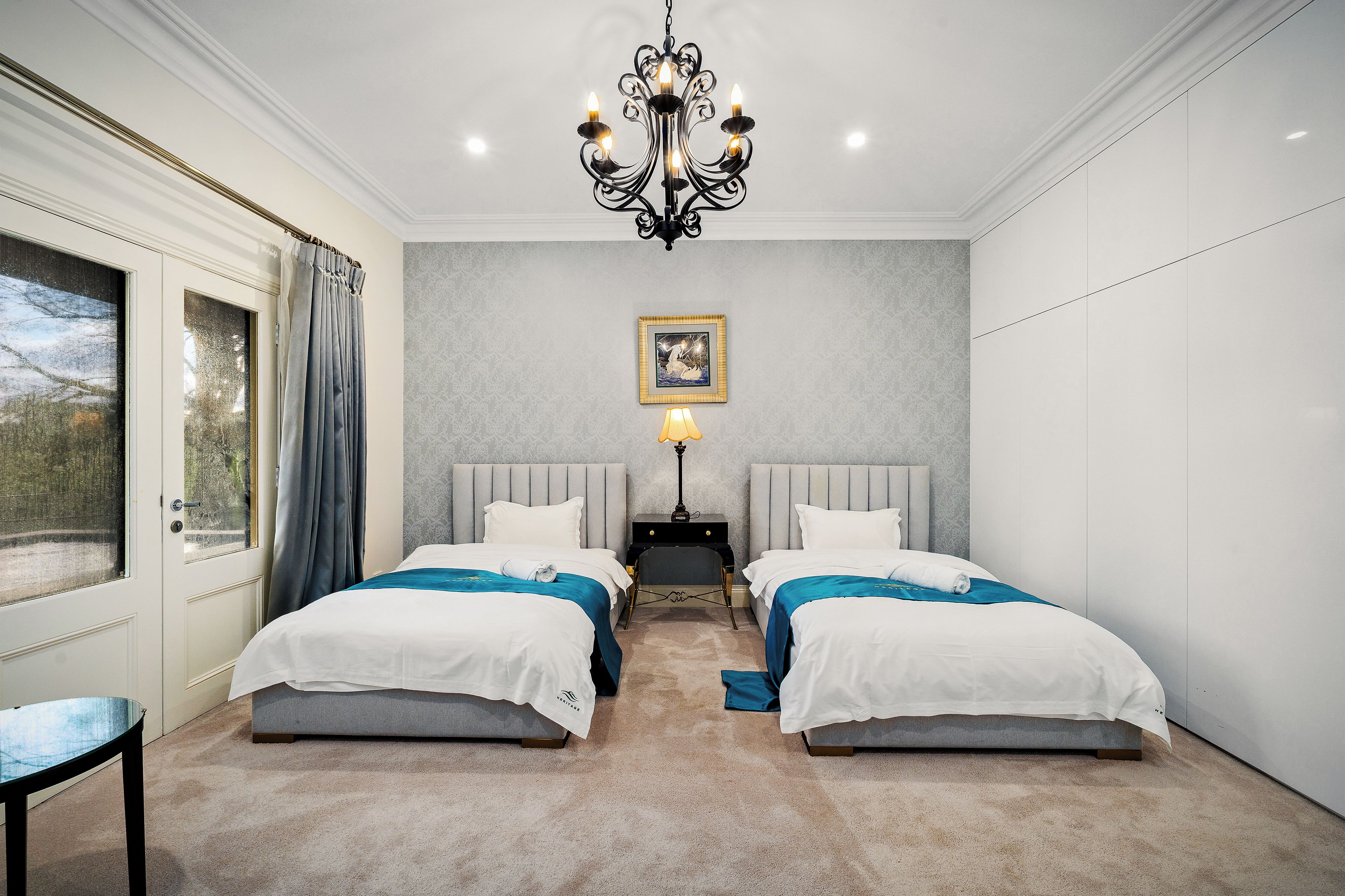 A hotel room with two beds, a chandelier, a nightstand, and a window with curtains.