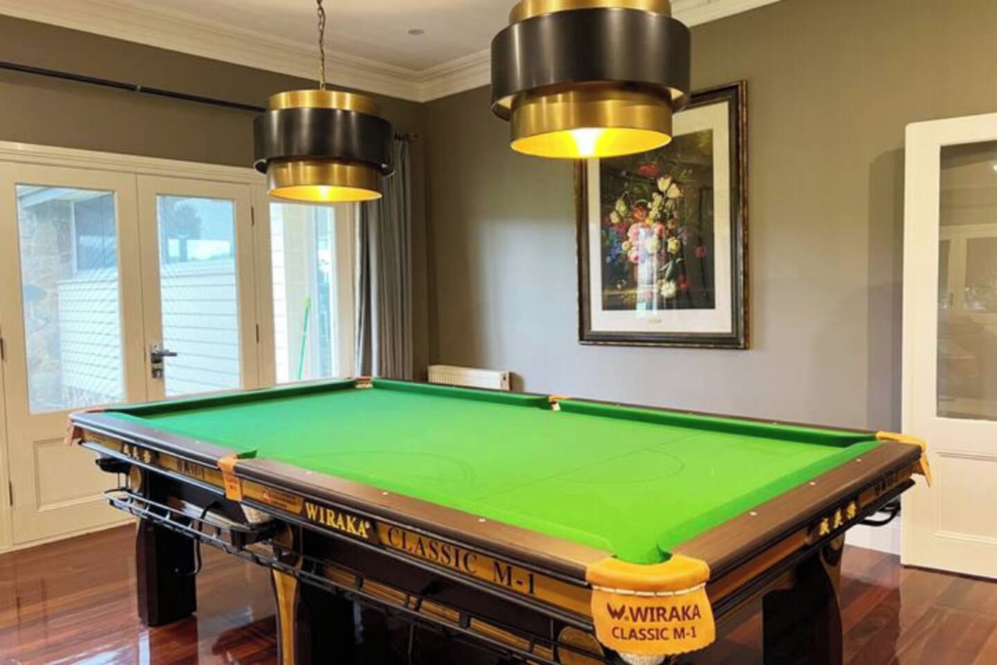 A pool table with a WIRAKA CLASSIC M-1 label, two hanging lamps, a framed floral painting, and a door with a view.