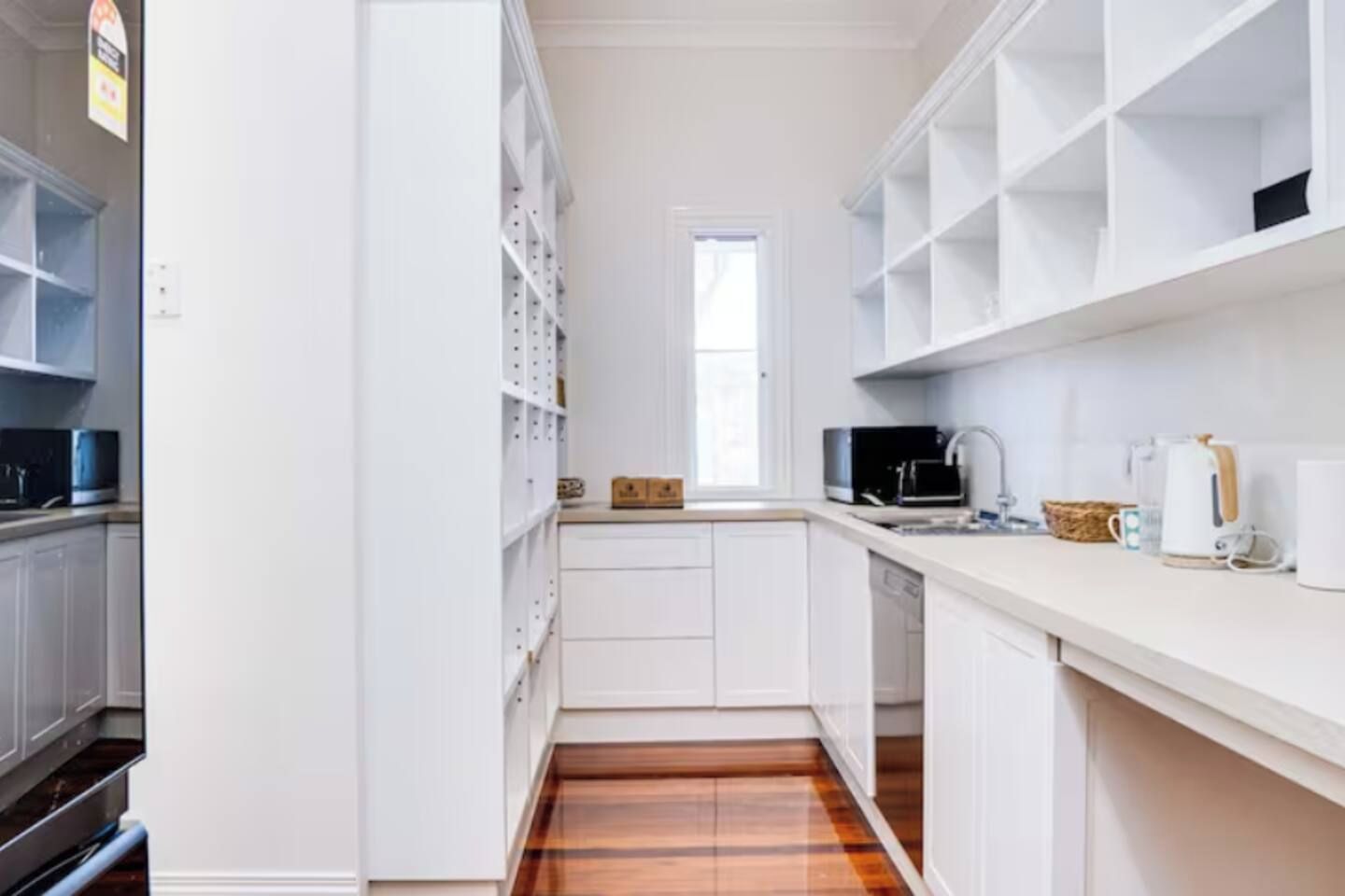 A narrow kitchen with white cabinetry, a wooden floor, and a window providing natural light.
