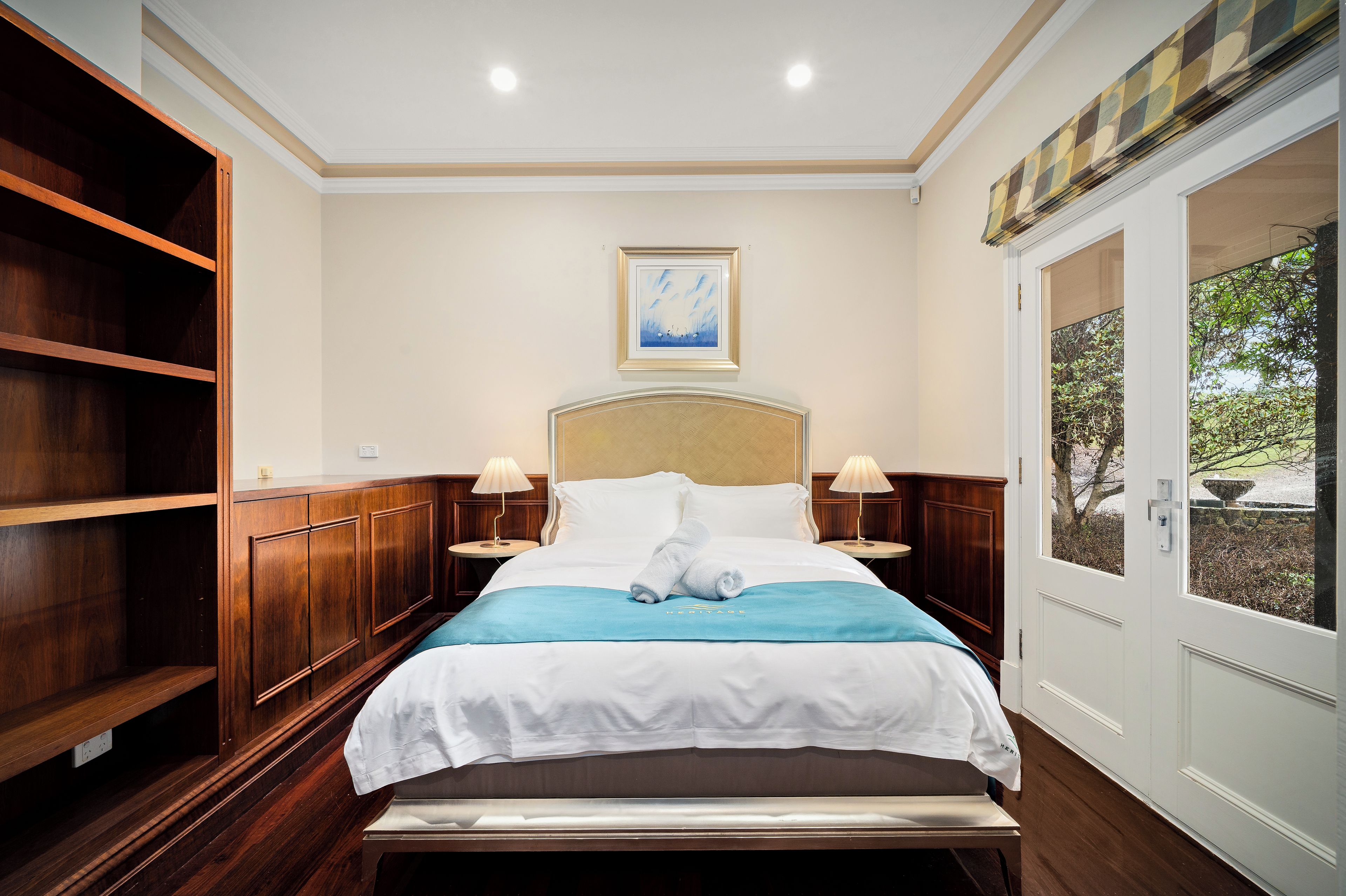 A hotel room with a bed, two bedside tables, a painting on the wall, and a view through a door.