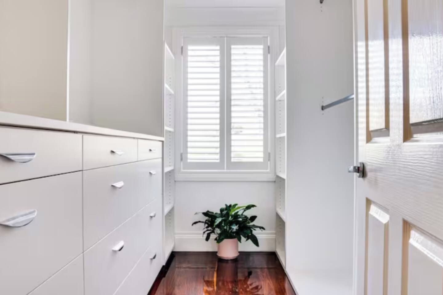 A narrow hallway with a white wardrobe, a potted plant, and a window with blinds.