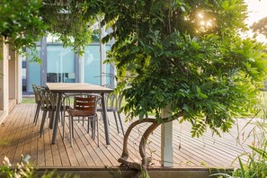 An outdoor wooden deck with a table and chairs, surrounded by greenery and a building in the background.