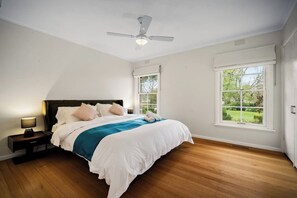 Un dormitorio con cama, mesita de noche, lámpara, ventilador de techo y ventanas grandes.