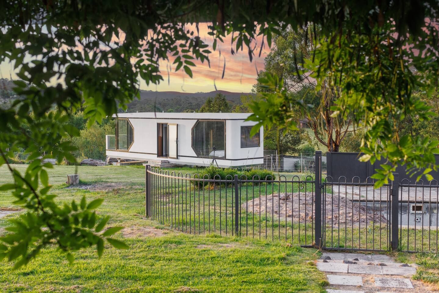 A modern house with a large glass window, surrounded by a metal fence, and set against a backdrop of trees and a distant landscape.