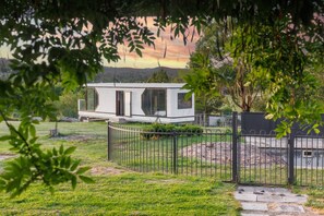 A modern house with a large glass window, surrounded by a metal fence, and set against a backdrop of trees and a distant landscape. - Yarra Valley Farmhouse | Alpacas Firepit Space Pod (Wonga Park)