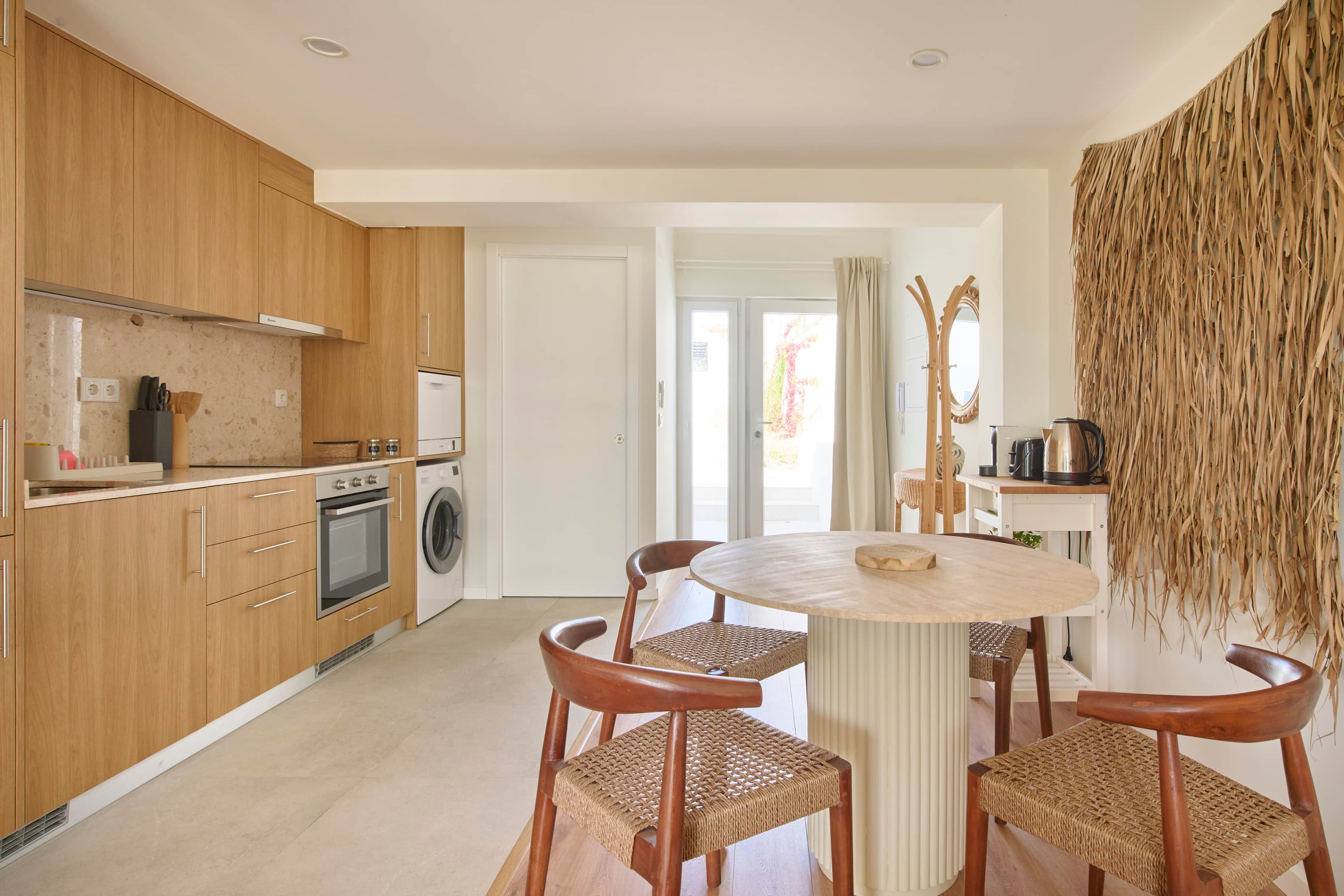 Cocina moderna con armarios de madera, horno empotrado y lavadora. Hay una mesa de comedor redonda con cuatro sillas de mimbre.