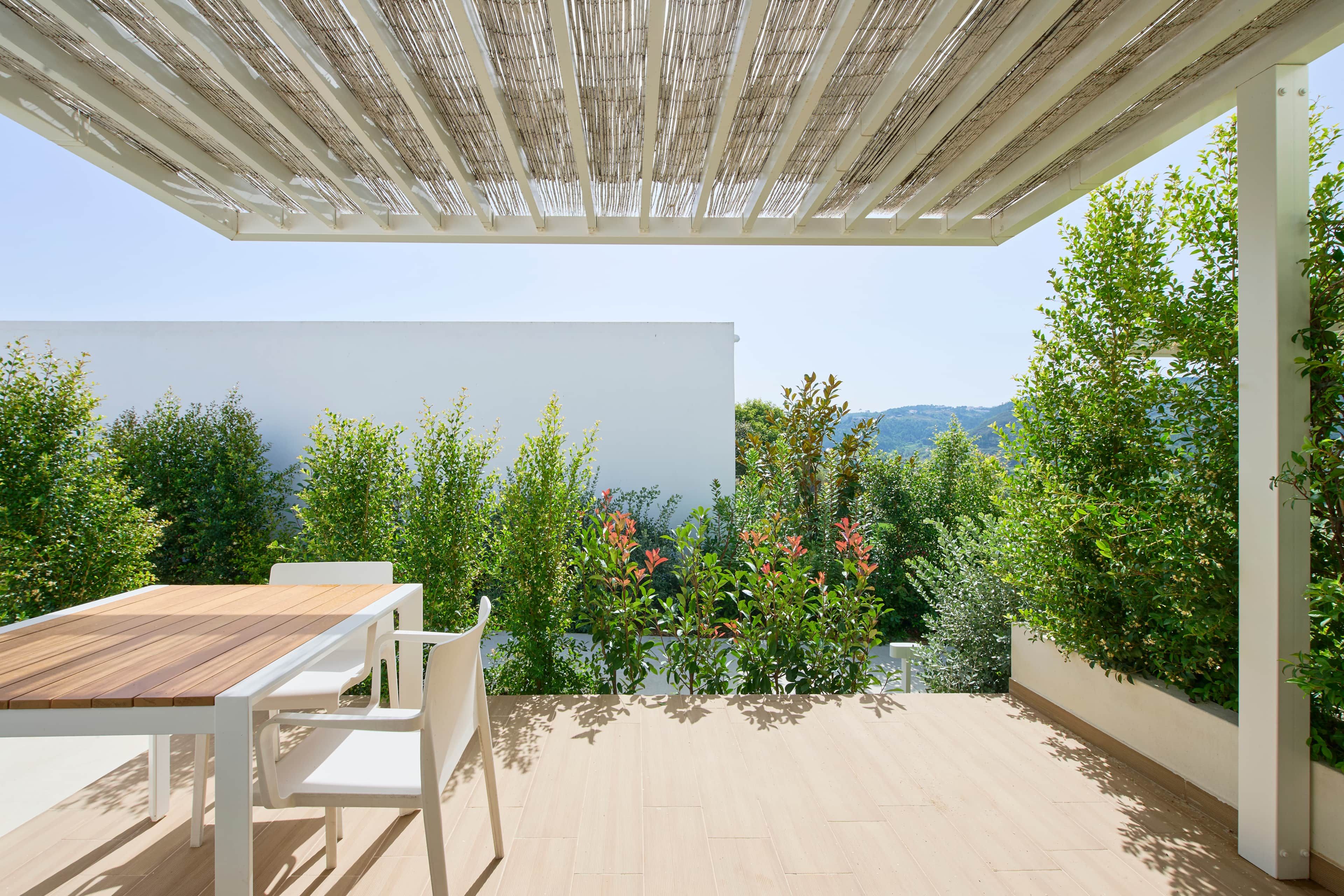 An outdoor patio with a wooden table and chairs, surrounded by greenery and a view of the landscape.