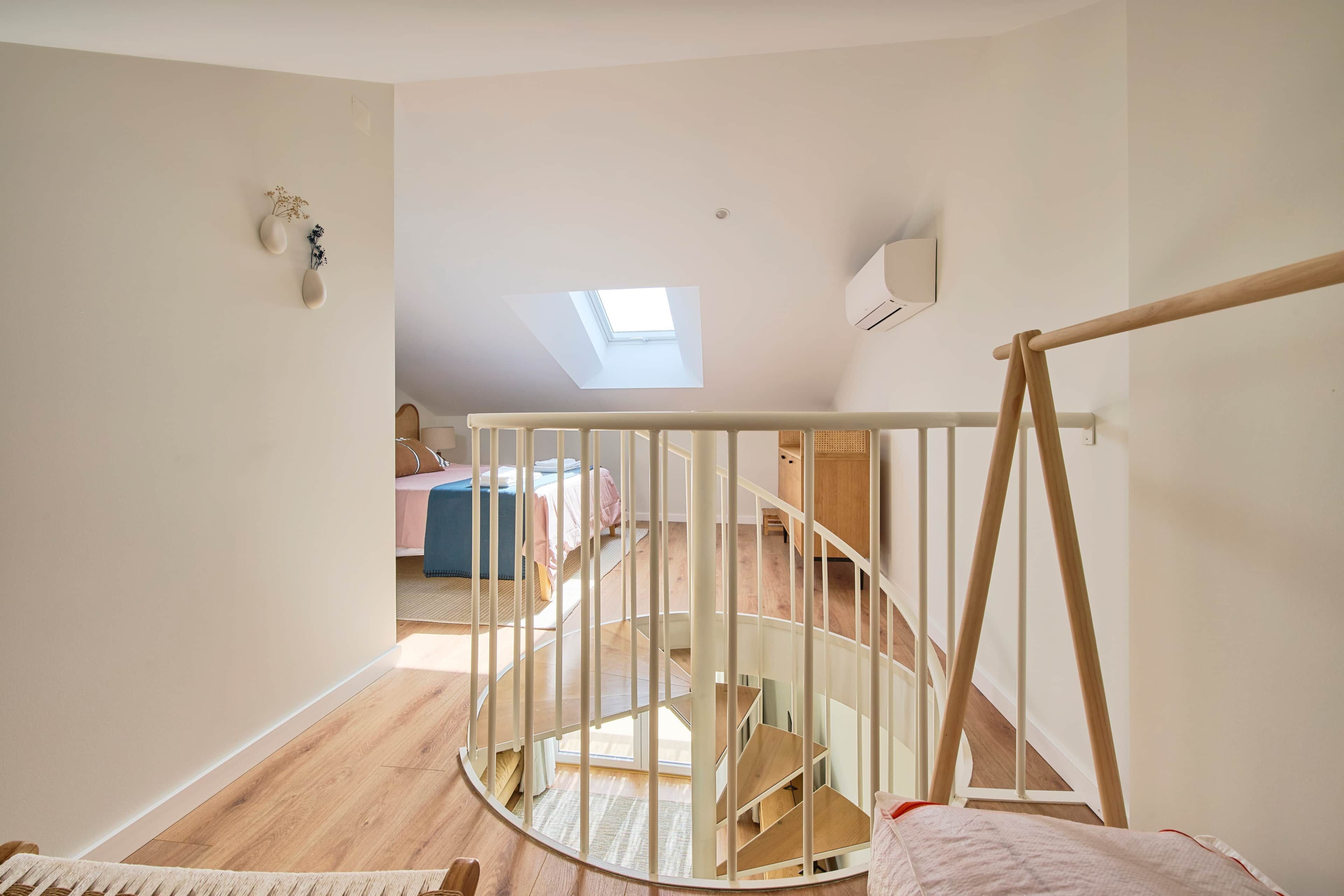 Een kamer met een houten trap, een dakraam en uitzicht op een andere kamer met een bed en bureau.