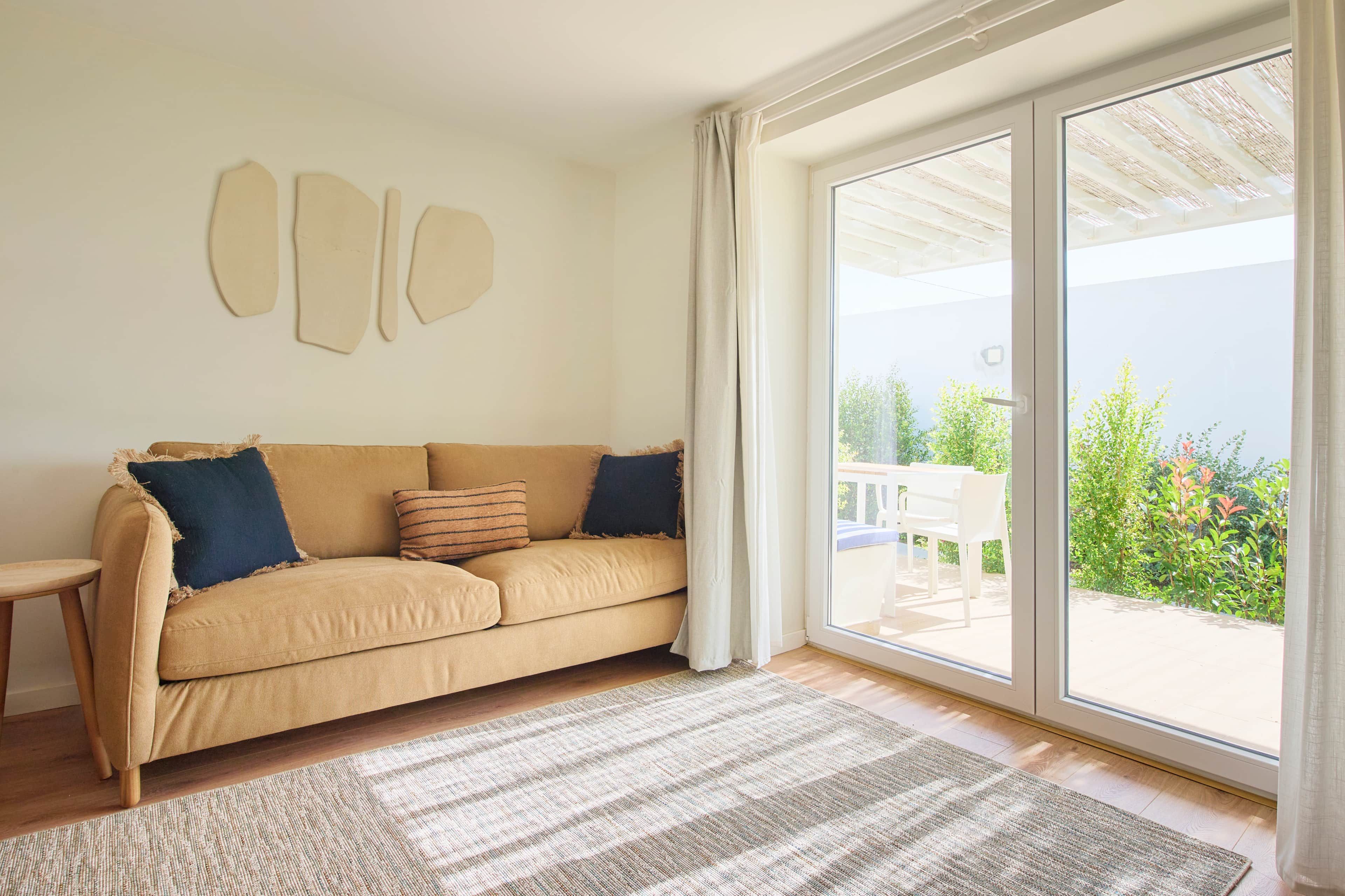 Un salón con sofá color beige, alfombra estampada y un amplio ventanal corredera que da a un espacio exterior con mesa y sillas.