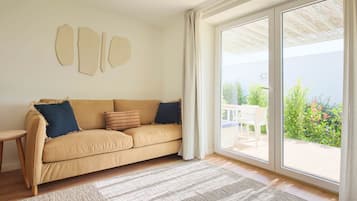 Un salón con sofá color beige, alfombra estampada y un amplio ventanal corredera que da a un espacio exterior con mesa y sillas.