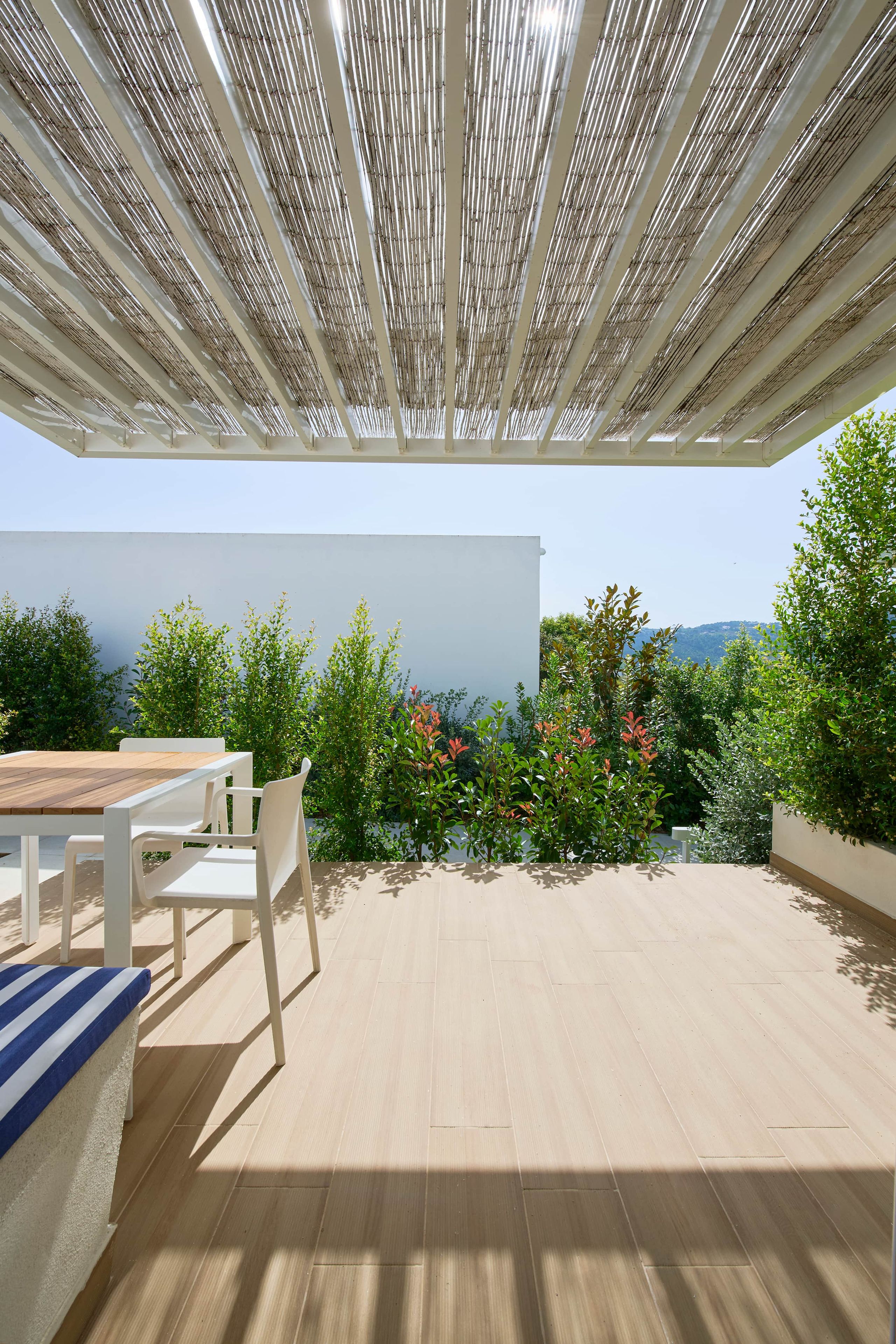 Un patio exterior con una mesa y sillas, una pérgola arriba y vistas a zonas verdes y colinas.