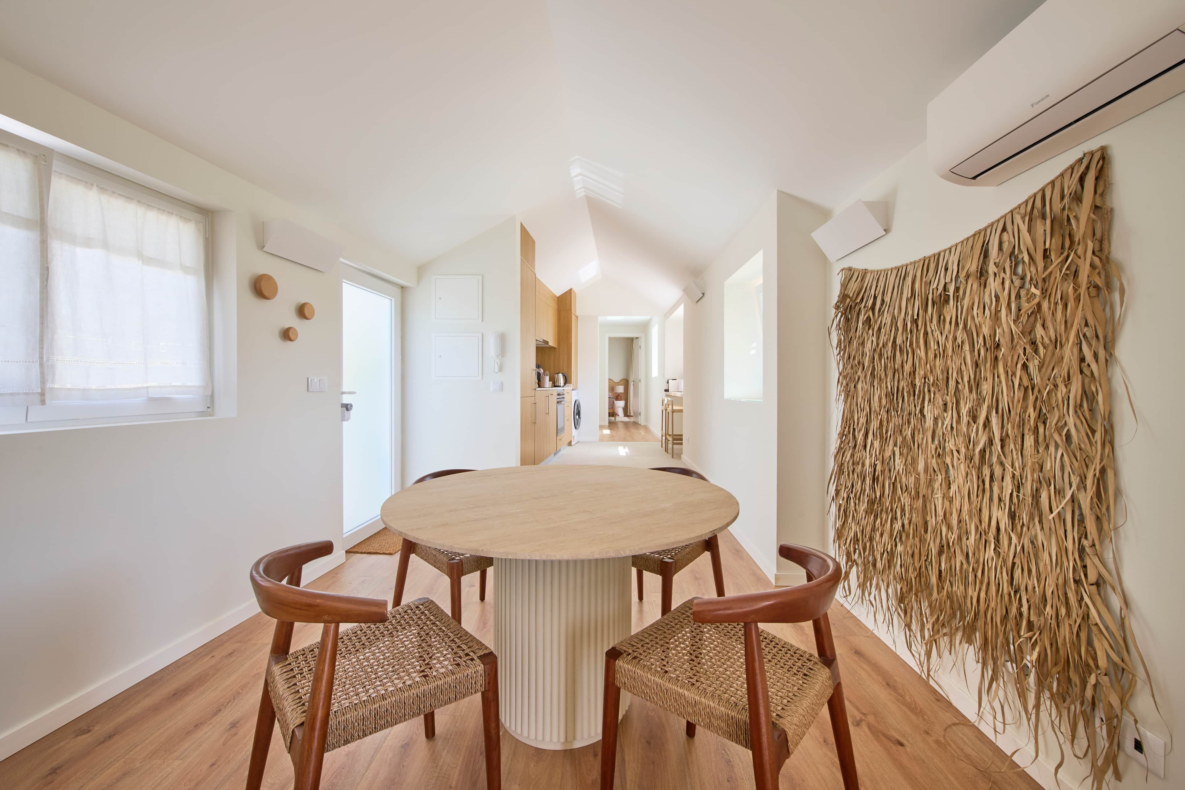 Una zona d'esmorzar moderna amb una taula rodona i quatre cadires, una cuina al fons i un penjoll decoratiu a la paret.