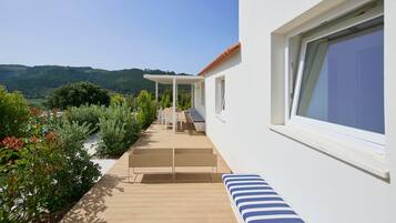 Un patio moderno al aire libre con terraza de madera, paredes blancas y vistas a colinas verdes.