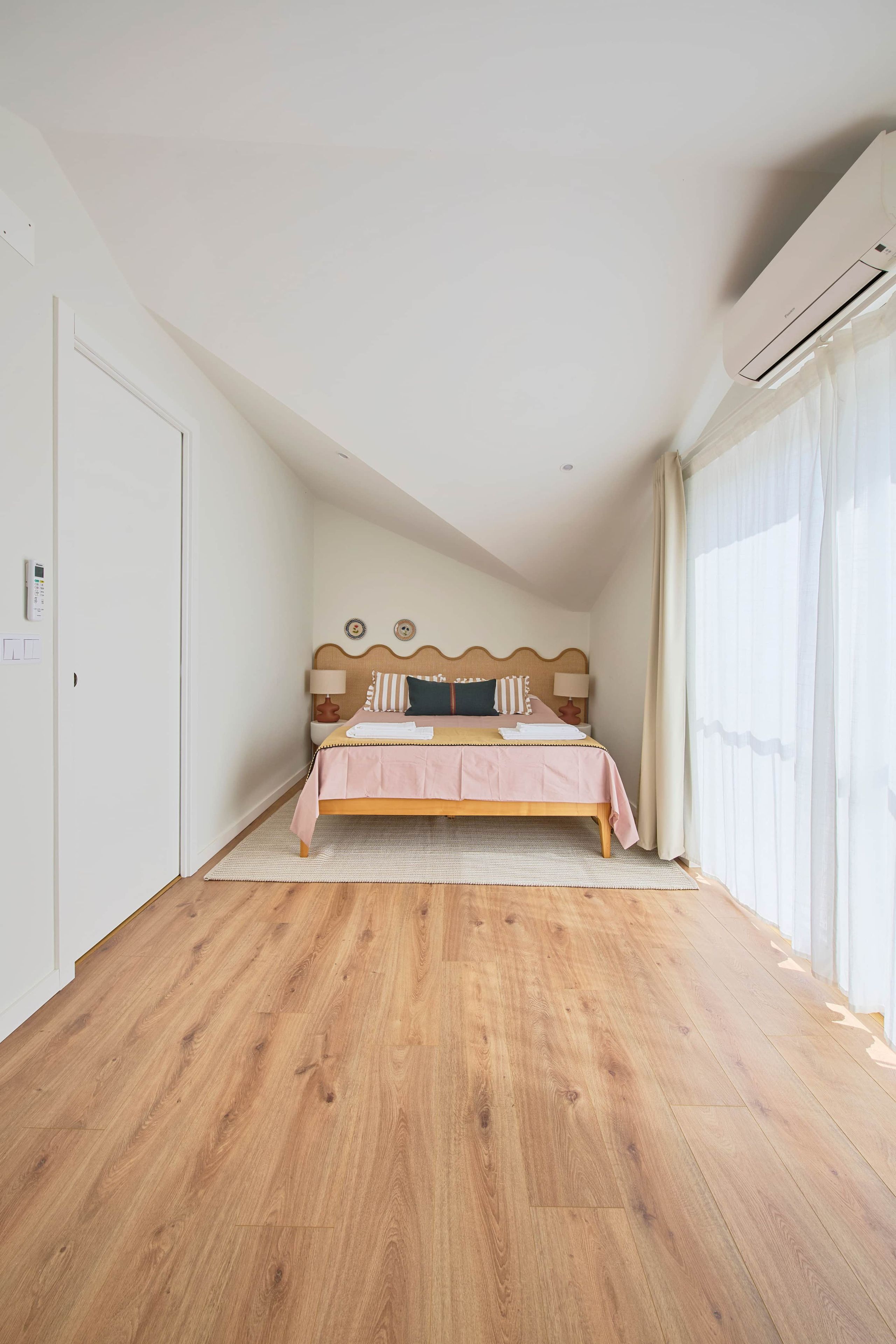 Un dormitorio con piso de madera, una cama con cubrecama rosa, un cabecero con detalle decorativo y una ventana con cortinas traslúcidas.