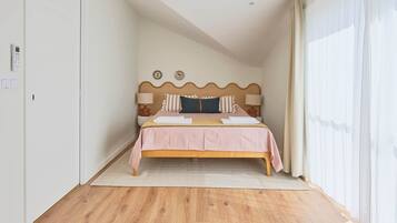 Un dormitorio con piso de madera, una cama con cubrecama rosa, un cabecero con detalle decorativo y una ventana con cortinas traslúcidas.