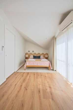 Une chambre avec un plancher en bois, un lit recouvert d’une couvre-lit rose, une tête de lit ornée d’un élément décoratif et une fenêtre agrémentée de rideaux légers.