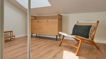 Una habitación con techo inclinado, piso de madera, un armario de madera, una silla de mimbre con cojín y un tragaluz.