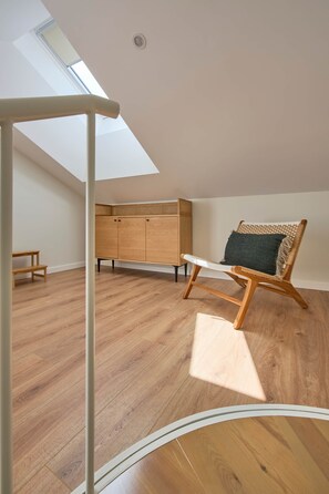 Een kamer met een schuine plafond, houten vloer, een houten kast, een rieten stoel met kussen, en een dakraam.
