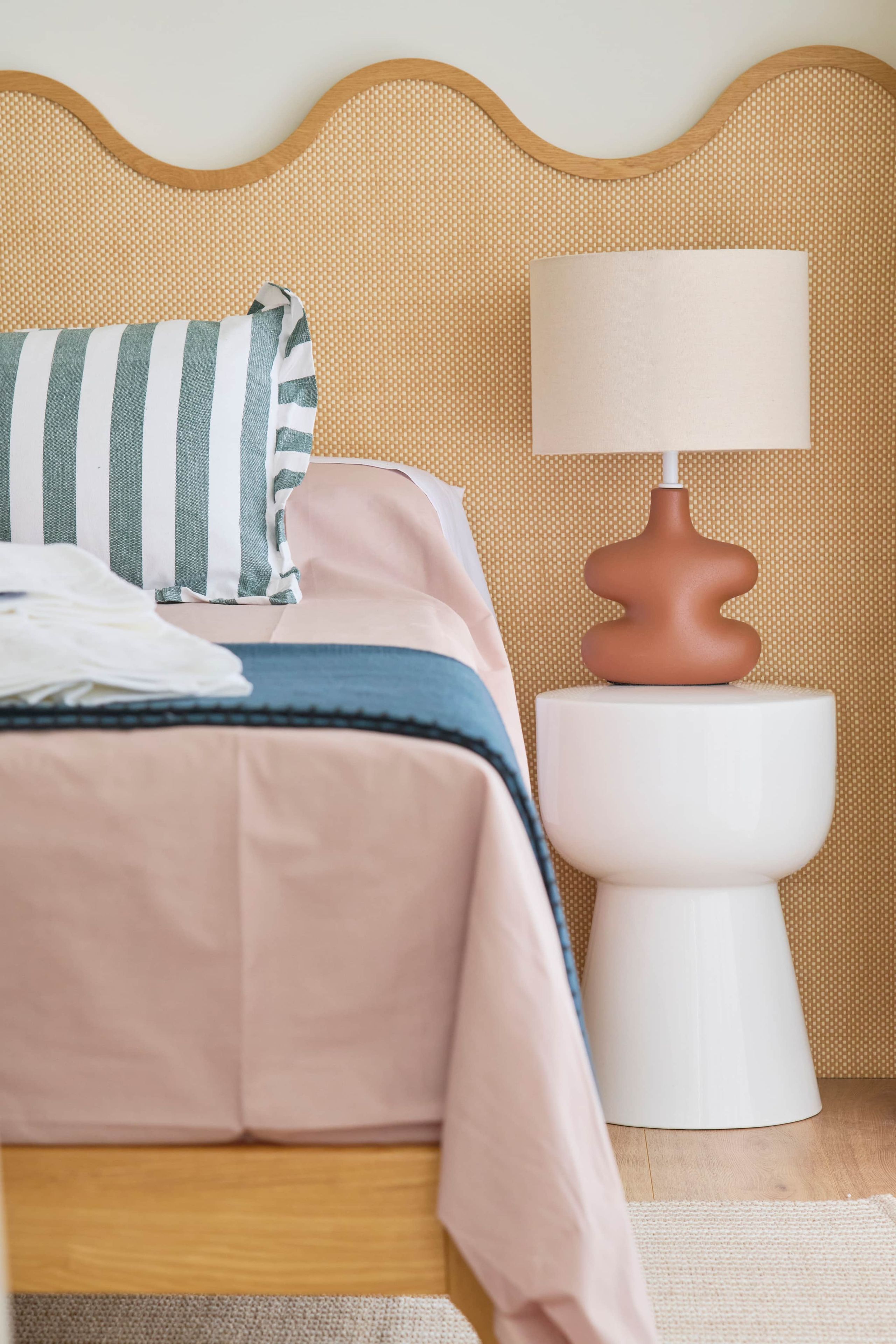 Una cama bien tendida con un almohadón a rayas y una mesita de noche blanca con una lámpara.