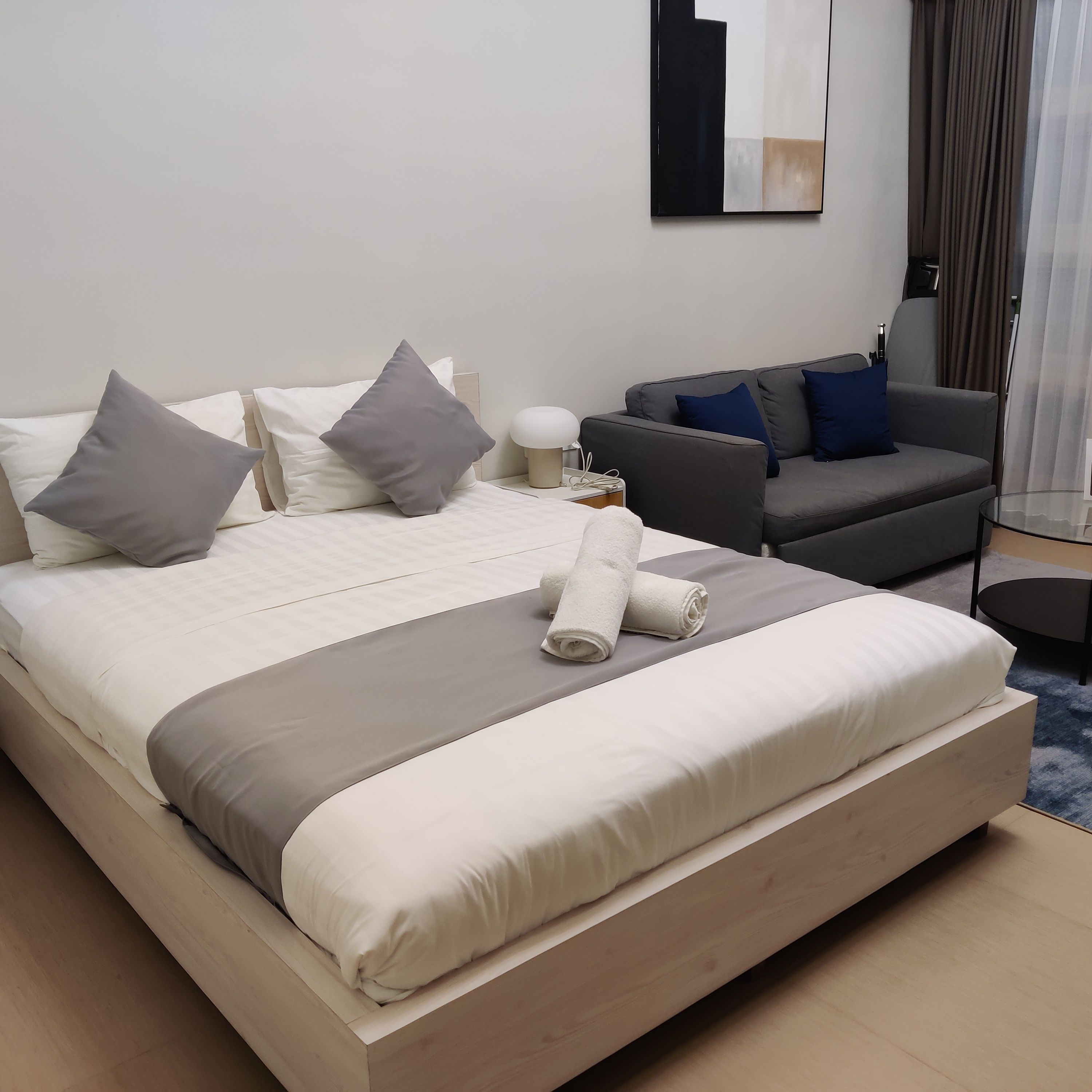 A modern hotel room with a large bed, a grey sofa, and a small side table.
