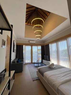 A modern hotel room with a large bed, a seating area with a sofa and armchairs, a dining table, and a view through large windows.