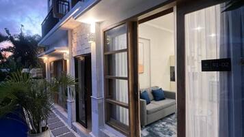 Un bâtiment moderne avec une entrée dotée d’une porte en verre, une pièce visible avec un canapé, et une enseigne portant l’inscription « R1-02 ».