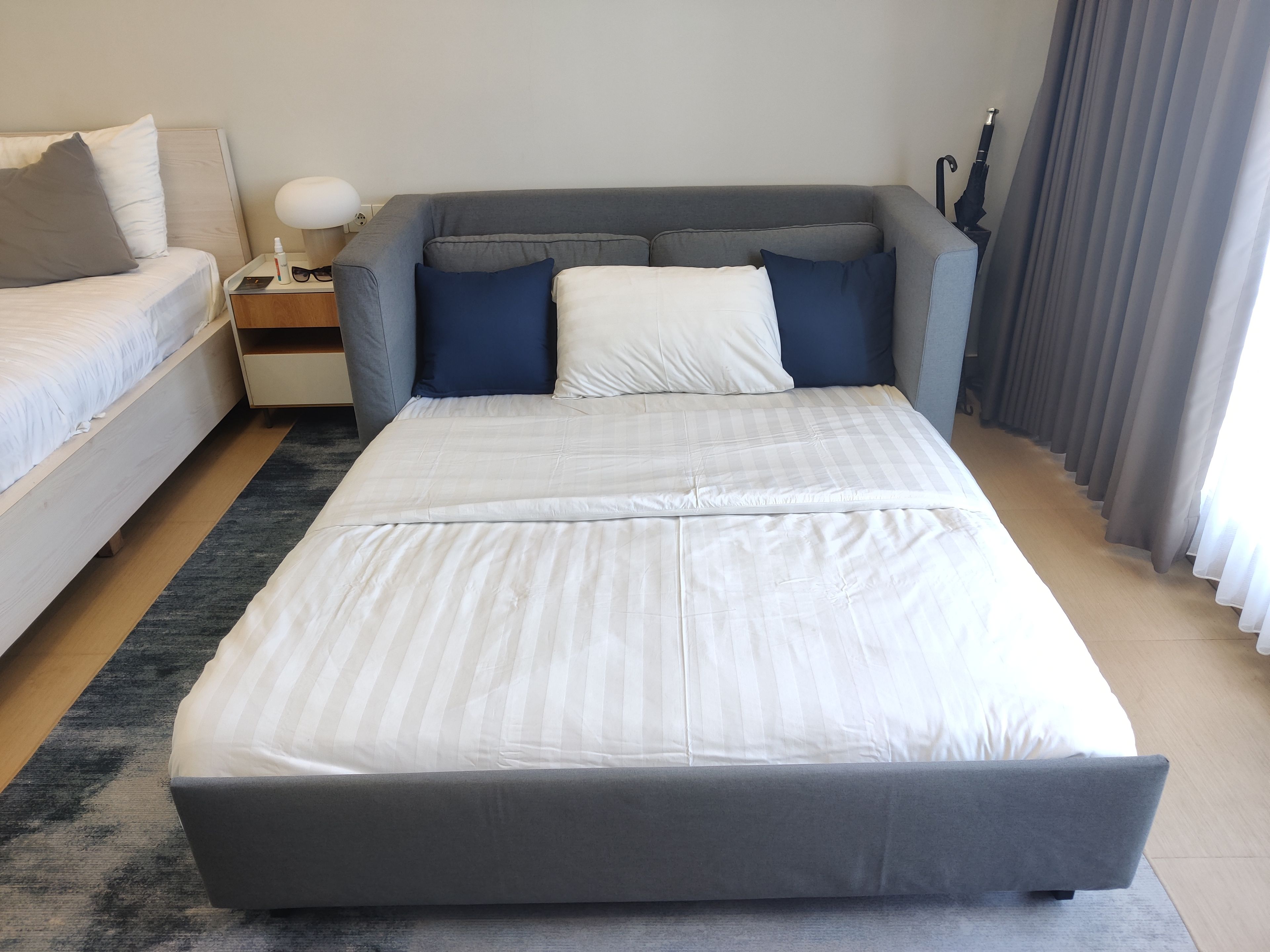 Una habitación de hotel moderna con una cama grande, una mesita de noche con lámpara y vistas al exterior.