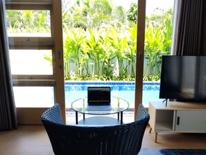 A room with a glass table, a laptop, a television, and a view of a swimming pool and greenery outside.