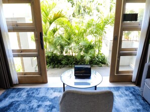 A room with a laptop on a glass table, a chair, and a view of a lush garden through open doors.