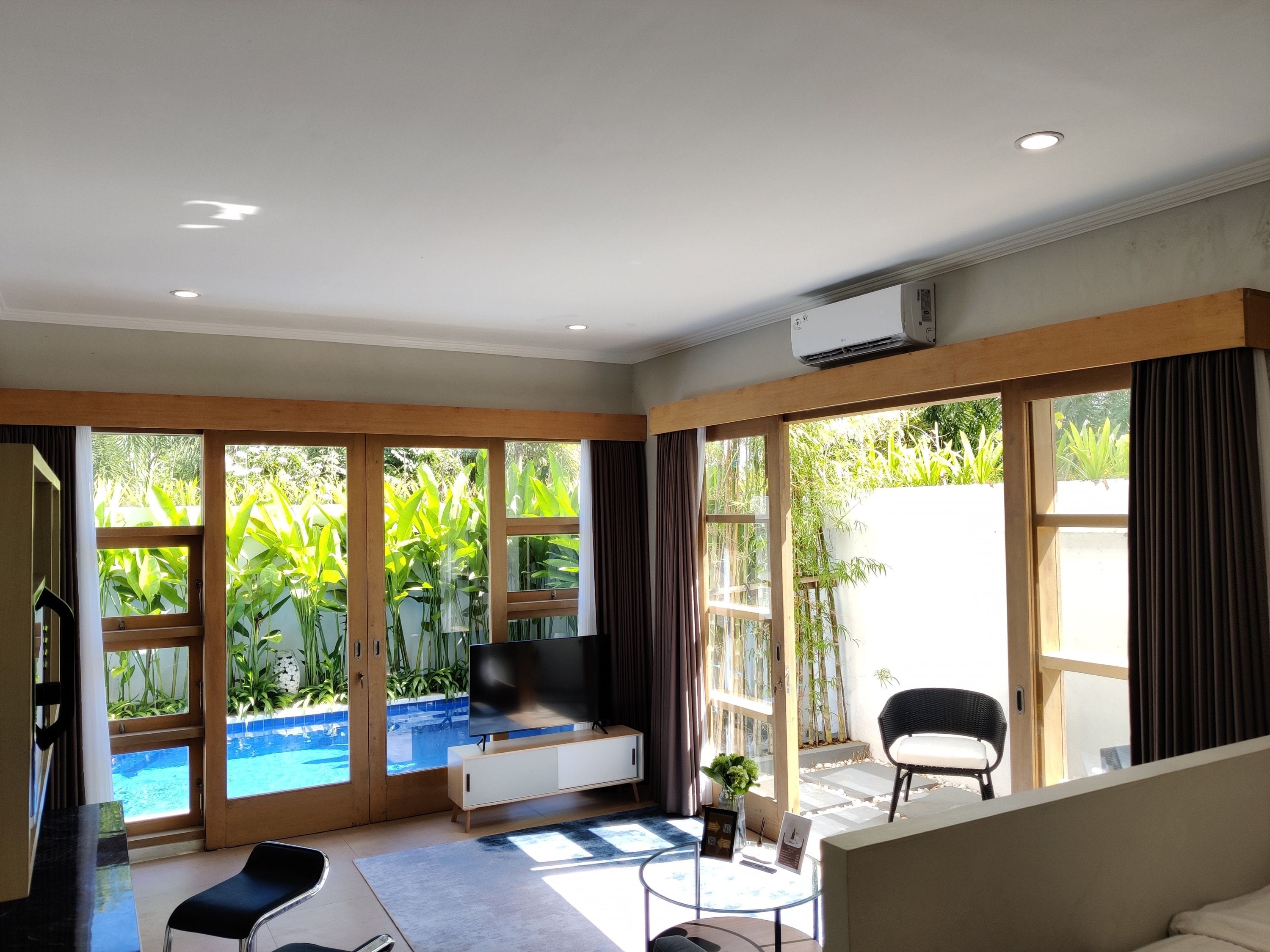 Ein geräumiges Wohnzimmer mit einem Flachbildfernseher, einer Couch, einem Couchtisch und großen Fenstern mit Blick auf einen Swimmingpool und Grünflächen.