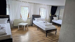 Una habitación con dos camas, una mesita pequeña y cortinas translúcidas en las ventanas.