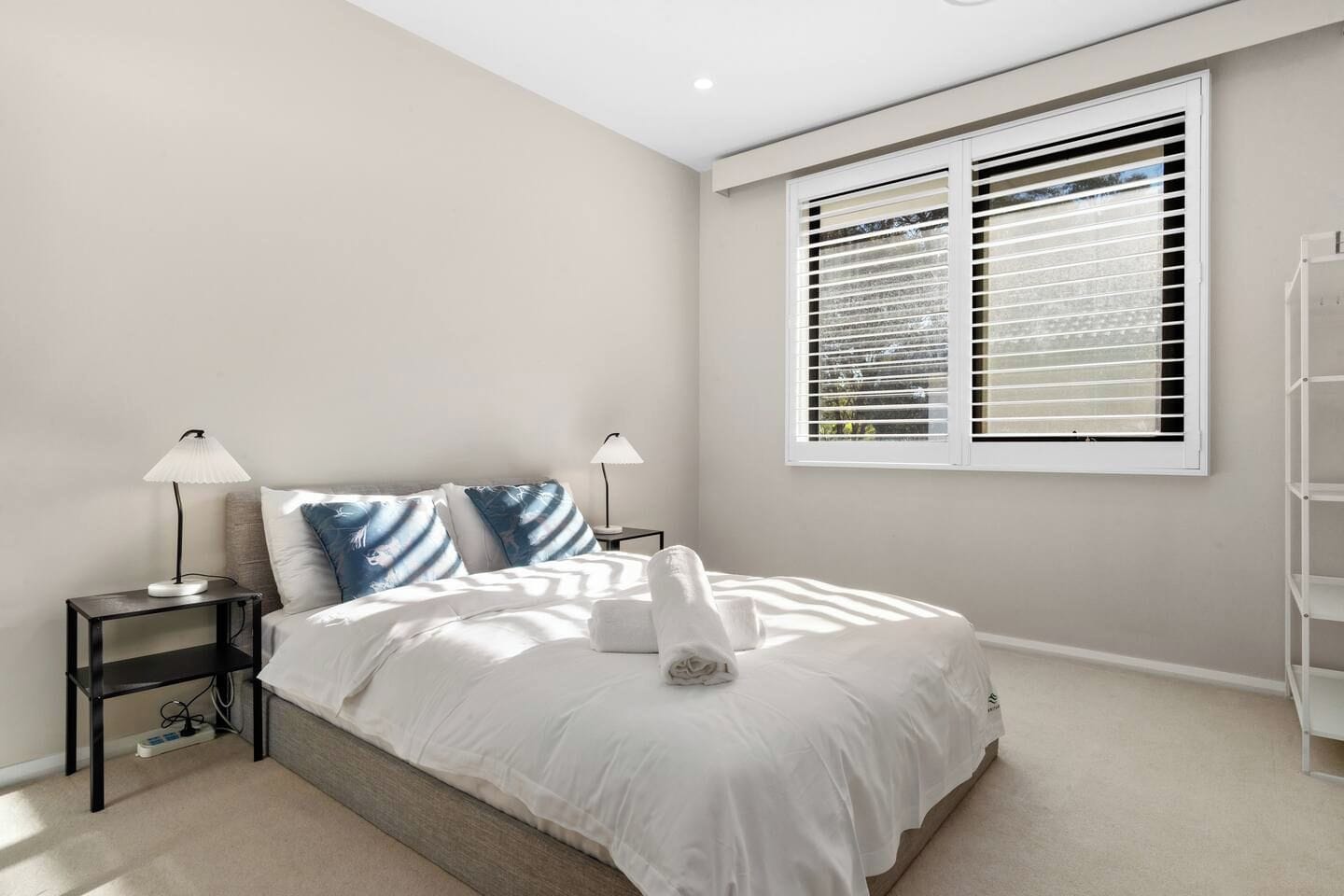 A bedroom with a bed, two bedside tables, a window with blinds, and a white wall.