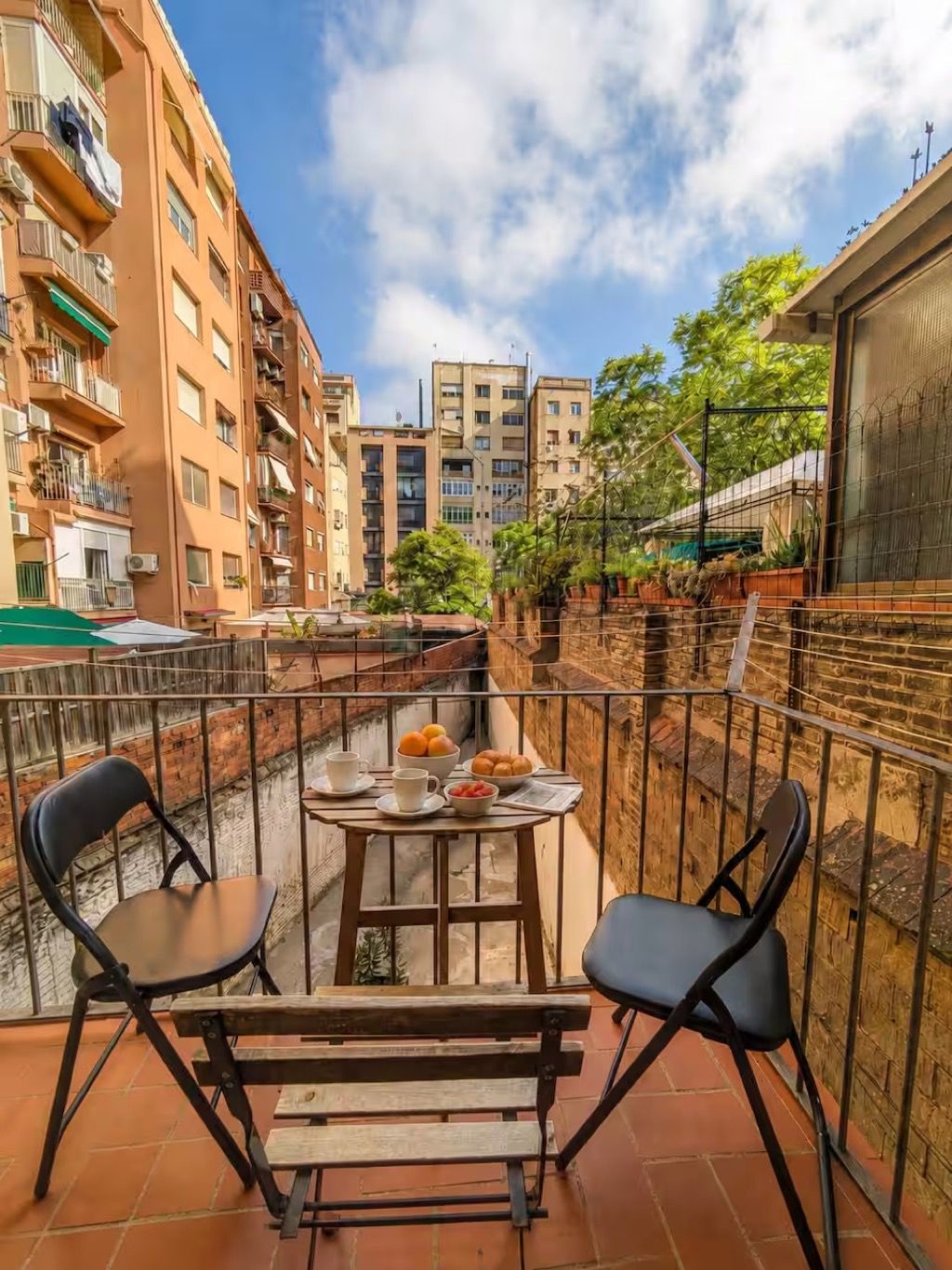 A balcony with two chairs and a table set for a meal, overlooking a narrow alley with buildings and a clear sky.