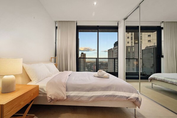 A hotel room with a bed, a nightstand, a lamp, a view of the city through large windows, and a balcony. - Skyline View, Crown Casino & Free Parking (Southbank)