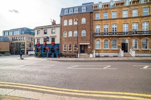 Straßenansicht mit Gebäuden, darunter ein Pub mit Blumenkästen, ein Backsteinhaus mit einem Schild und ein modernes Gebäude.